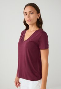 Donna con i capelli castani fino alle spalle, che indossa una maglietta a maniche corte di colore bordeaux con scollatura a V e pantaloni bianchi, in piedi contro uno sfondo chiaro e uniforme.
