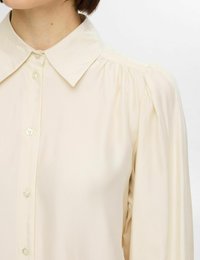 Krämfärgad blus med klassisk krage, knappstängning fram, samlade axlar och slät, blank tygstruktur.