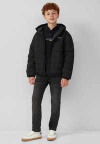 Zwarte pufferjack met een capuchon, voorzien van een rits en ribgebreide manchetten, gedragen over een zwarte hoodie. Gecombineerd met donkere jeans en witte sneakers.
