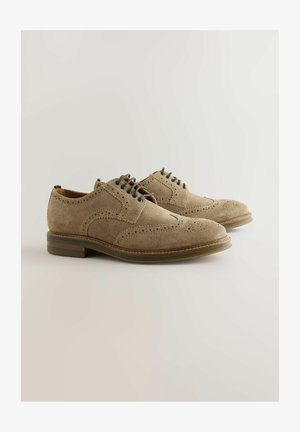 Brogue kurpes no dzeltenas zamšas ar uzspraužamiem spārniņiem un brūniem gumijas zolēm uz gaiša fona.