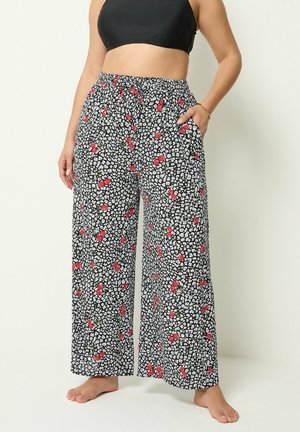 Kvinde iført sorte bukser med vide ben med hvidt og rødt kirsebærmønster og sort crop top, stående barfodet med den ene hånd i lommen.