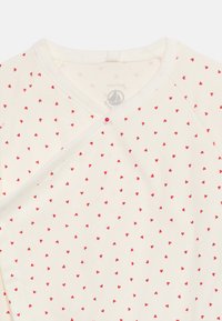 Κρεμ χρώματος μωρουδιακό πουκάμισο τύλιγμα με μικρά κόκκινα σχέδια καρδιάς και κόκκινο κουμπί κοντά στο λαιμό.