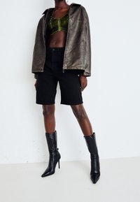 Černá kožená bunda, zelený zkrácený top, černé džíny šortky a černé lodičky na vysokém podpatku s žebrovanou texturou a špičatými špičkami, na neutrálním pozadí.