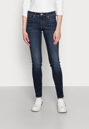 Dark denim jeans with a slim fit, featuring a classic five-pocket design, brass button closure, and contrasting stitching. Worn with white sneakers.