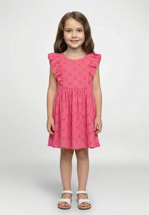 Robe en coton rose avec motifs en œillets, épaules à volants et jupe évasée. Associée à des sandales blanches.