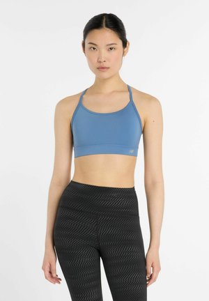 Kvinde iført en blå sports-bh og sorte leggings med høj talje og mønster, stående mod en ensfarvet hvid baggrund.