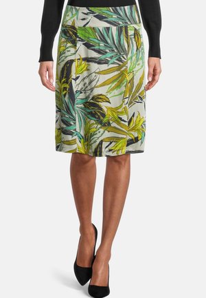 Jupe mi-longue avec un motif de feuilles tropicales multicolores en vert, jaune et noir, portée avec un haut à manches longues noir et des talons noirs.