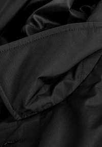 Gros plan sur un tissu noir, présentant une texture lisse avec un léger éclat. Le matériau est léger, avec des coutures visibles et des plis.