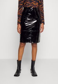 Jupe crayon en cuir verni noir avec taille haute, coutures avant et finition brillante. Porte avec un haut à manches longues à motifs et des bottines.