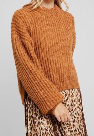 Ein rostfarbener, gerippter Strickpullover mit hohem Kragen und weiten, oversized Ärmeln, kombiniert mit einem Rock im Leopardenmuster.