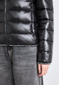 Svart dunjakke med glansfull finish, med glidelås og unik Tommy Jeans-merket elastisk kant. Grå denimbukser synlige nedenfor.