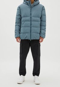 Polstret teal-jacka med huva, med en framdragkedja, vertikala sömmar och en lätt glänsande yta. Matchas med svarta cargobyxor och vita sneakers.