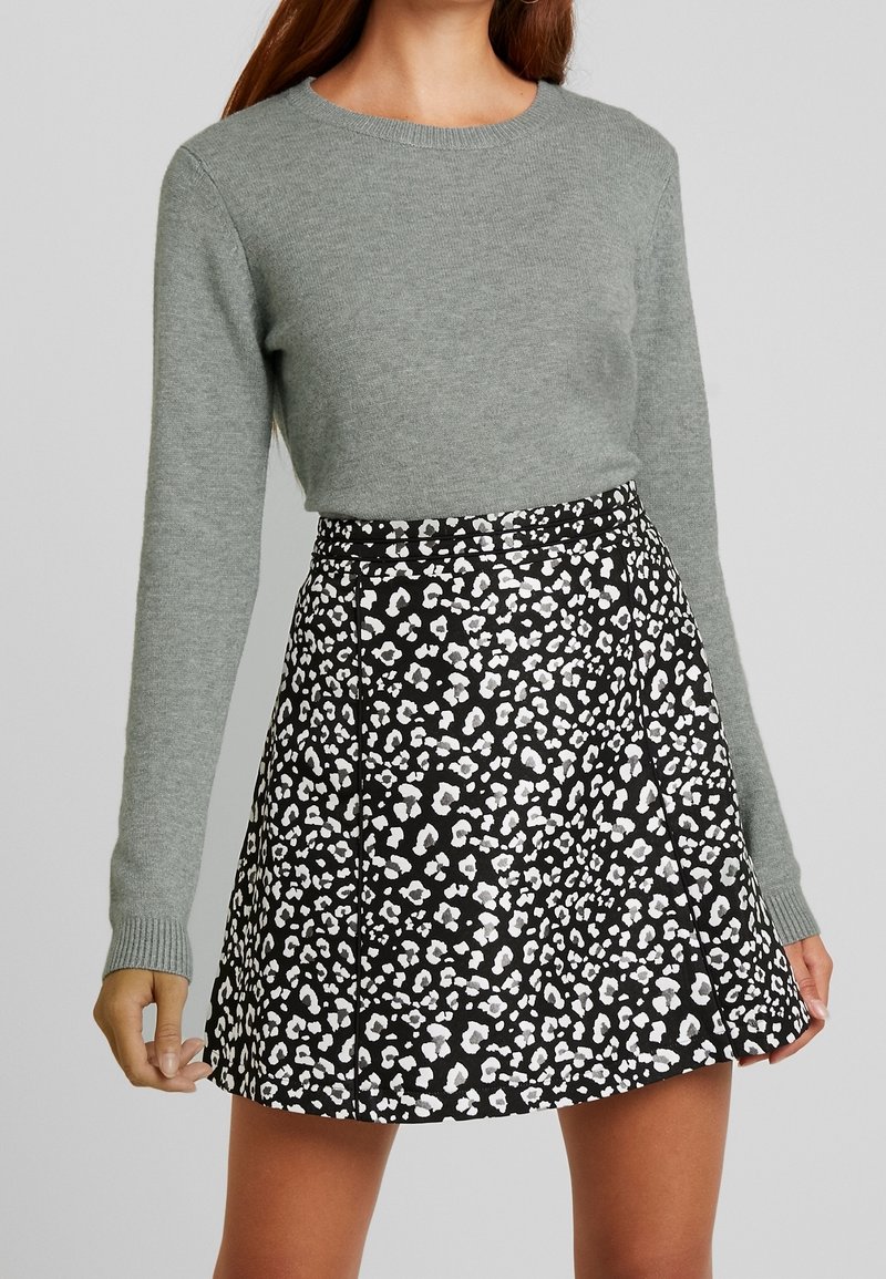 Femme portant un pull gris à manches longues et une jupe noire trapèze avec un motif léopard blanc et gris clair.