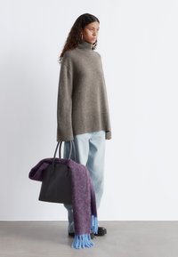 Übergroßer, grauer Rollkragenpullover mit geripptem Saum, kombiniert mit hellblauen Jeans. Schwarze Tragetasche mit lila und blauer Fransen-Schal, der darüber drapiert ist.