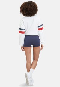 Witte cropped sweatshirt met rode en marineblauwe strepen op de mouwen, gecombineerd met marineblauwe shorts. Model draagt witte sneakers en enkelsokken.