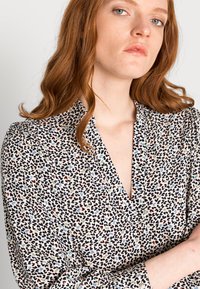 Bloemendessinf blouse van lichte stof; heeft zwarte, rode, blauwe en groene accenten op een crèmekleurige achtergrond; V-hals en knoopsluitingen bij de mouwen.