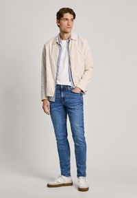 Chaqueta beige claro sobre una camiseta blanca, combinada con jeans azules y zapatillas blancas. La chaqueta tiene un diseño simple y un corte ajustado.