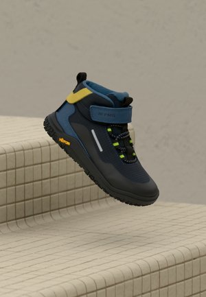 Granatowo-czarny sneaker wysokiej góry z żółtym paskiem, odblaskowym detalem i podeszwą Vibram, umieszczony nad beżowymi, wyłożonymi kafelkami stopniami.