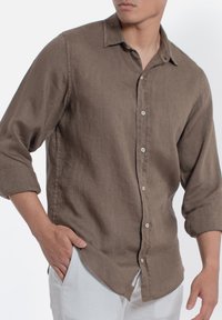 Camicia button-up in lino marrone con colletto e maniche arrotolate, caratterizzata da un orlo dritto e una texture sottile. Indossata con pantaloni chiari.