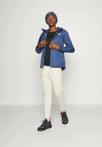 Person som har på seg blå lue, blå jakke, svart skjorte, hvite bukser og svarte joggesko med rosa detaljer, står mot en enkel lys bakgrunn.