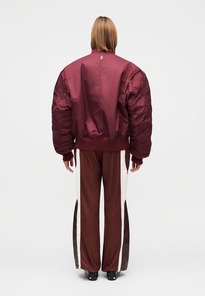 Veste matelassée bordeaux avec manches surdimensionnées et poignets côtelés, associée à un pantalon marron avec des bandes latérales crème et marron foncé.