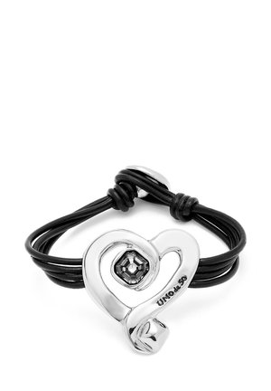 Charm plateado en forma de corazón con un cristal negro central, montado en cuerdas de cuero negro, con un diseño en bucle y grabado de firma.