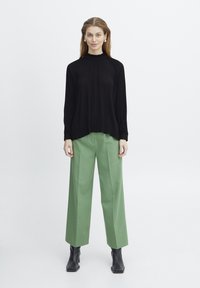 Haut noir à manches longues côtelé avec un col montant, associé à un pantalon vert ample et des bottines noires.