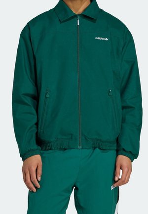 Homme portant une veste zippée Adidas vert foncé avec poignets élastiques et pantalon assorti, mains détendues sur les côtés.