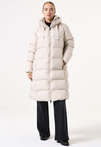 Abrigo tipo puffer beige con diseño acolchado, capucha y cremallera frontal, combinado con pantalones anchos negros y zapatos negros.
