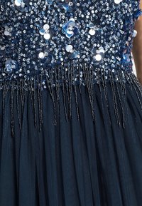 Vestido azul marino con un torso de lentejuelas con adornos de lentejuelas en forma circular y detalle de flecos verticales, que fluye hacia una falda de tul texturizado.