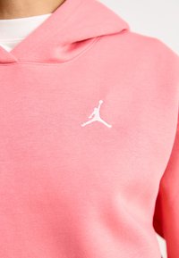 Ružová mikina z mäkkej látky, s bielym logom Jumpman na hrudi. Dizajn obsahuje klokanie vrecko a kapucňu so šnúrkou.