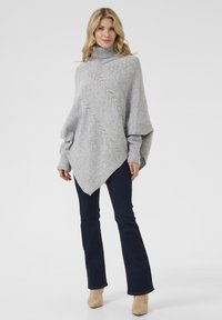 Pelēks adīts pončo ar augstu apkakli, ar zigzaga rakstu un asimetrisku apakšmalu, apvienots ar tumši zilu platu džinsu biksēs un bēšiem potīšu zābakiem.