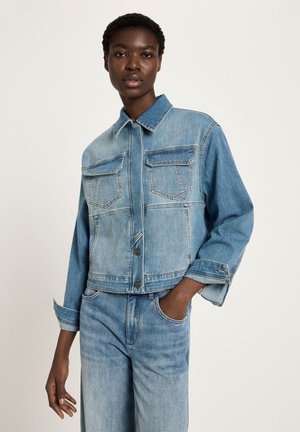 Frau trägt eine hellblaue Jeansjacke mit Klappentaschen und passende, hoch taillierte Jeans, eine Hand in der Tasche, neutraler Hintergrund.