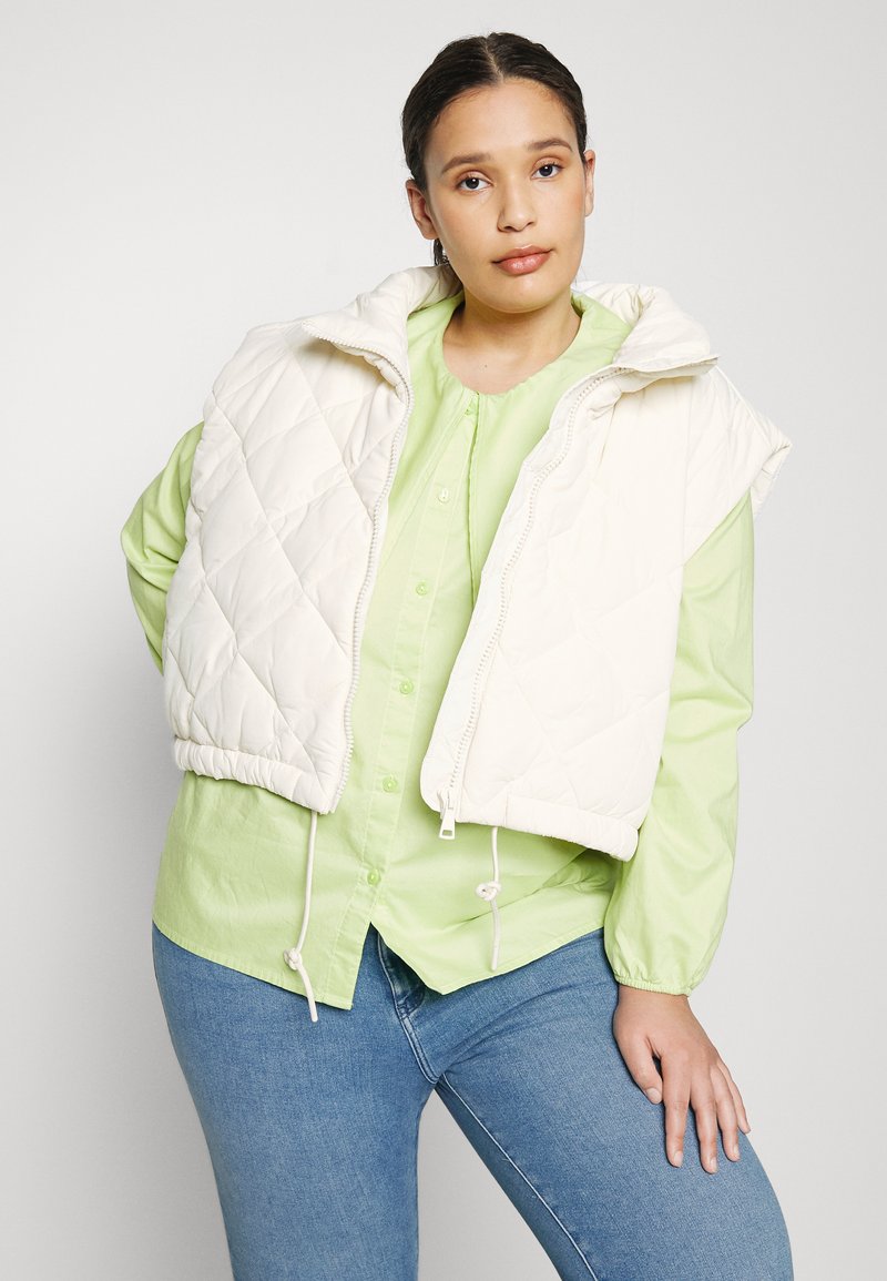 Kremowa pikowana kamizelka z wysokim kołnierzem, zamkiem błyskawicznym z przodu i ściągaczem na dole, noszona na jasnoszarej koszuli na guziki. Ciemnoniebieskie dżinsy.