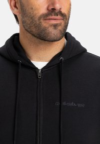 Sweatshirt preto com fecho éclair feito de tecido suave, apresentando um capuz com cordão e um logótipo bordado discreto no peito. Gola moderna e detalhes de costura.