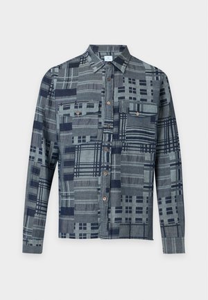 Jeune homme portant une chemise grise à motifs boutonnée, un pantalon beige à jambes larges et des chaussures marron bicolores, debout devant un fond blanc.