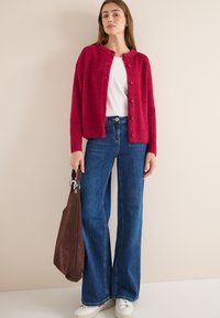 Rote, flauschige Strickjacke mit Rundhalsausschnitt und Knöpfen, kombiniert mit hoch taillierten, weitgeschnittenen blauen Jeans und einer braunen Wildledertasche. Weiße Sneakers werden getragen.