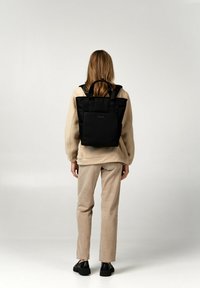 Zaino tote in tessuto nero con doppi manici superiori, forma rettangolare e tasche laterali esterne, indossato sopra un outfit beige chiaro.