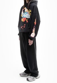 Sudadera negra con gráficos coloridos que incluyen corazones y arcoíris, combinada con pantalones holgados negros y zapatillas metálicas.