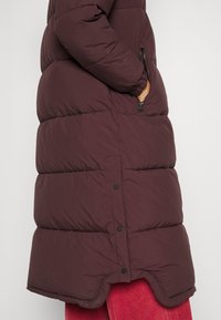 Bordeauxfarbener Puffer-Mantel mit einem gesteppten Design, seitlichen Reißverschlusstaschen und einem abgerundeten Saum mit Druckknopfverschlüssen.