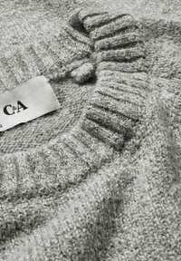 Pull en tricot gris avec col côtelé, présentant une finition texturée et une étiquette marquée "C&A". Matière douce avec des détails de couture visibles.
