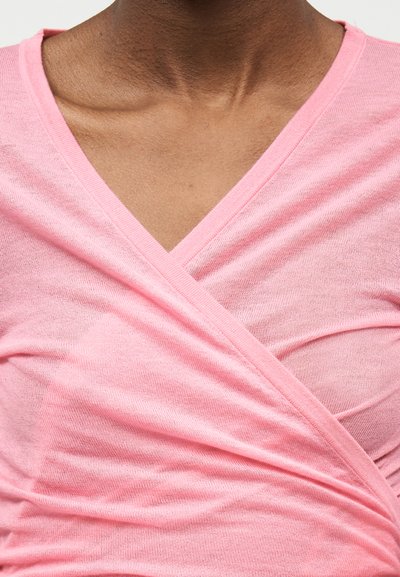 Personne à la peau foncée portant un haut cache-cœur rose pâle à col en V, en tricot fin avec des plis subtils sur la poitrine et les épaules.