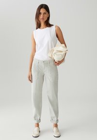 Top senza maniche bianco abbinato a pantaloni grigi chiari con tasche anteriori e orli risvoltati. Il modello tiene una borsa beige. Colori solidi, design informale.