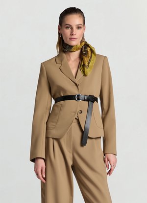 Femme portant un costume beige avec une ceinture noire autour de la veste et une écharpe verte à motifs nouée autour du cou.