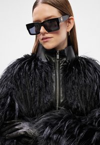 Casaco de pelo sintético preto com colarinho e punhos em couro, combinado com óculos de sol oversized pretos com detalhes em hardware dourado.