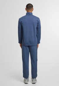 Tumši zilā flīsa sporta uzvalks ar augstu apkakli, elastīgiem maliņām un jostu. Biksēm ir sānu kabatas, kopā ar vieglām sporta apavām.