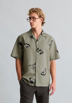 Jeune homme aux cheveux blonds et lunettes, portant une chemise verte à manches courtes avec des motifs noirs de fruits de mer et d'agrumes, debout devant un fond uni.