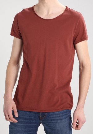 Uomo con indosso una maglietta a maniche corte color ruggine semplice e jeans blu, ripreso dal collo ai fianchi su uno sfondo uniforme.