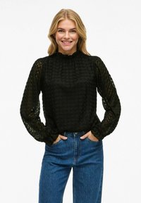 Blusa nera con texture, maniche lunghe in chiffon, collo alto e polsini arricciati, abbinata a jeans blu a vita alta.