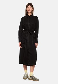 Schwarzes Hemdkleid mit langen Ärmeln, Knopfleiste vorne, Gürtel in der Taille und kontrastierenden Nähten. Kombiniert mit gemusterten Turnschuhen und gelben Socken.